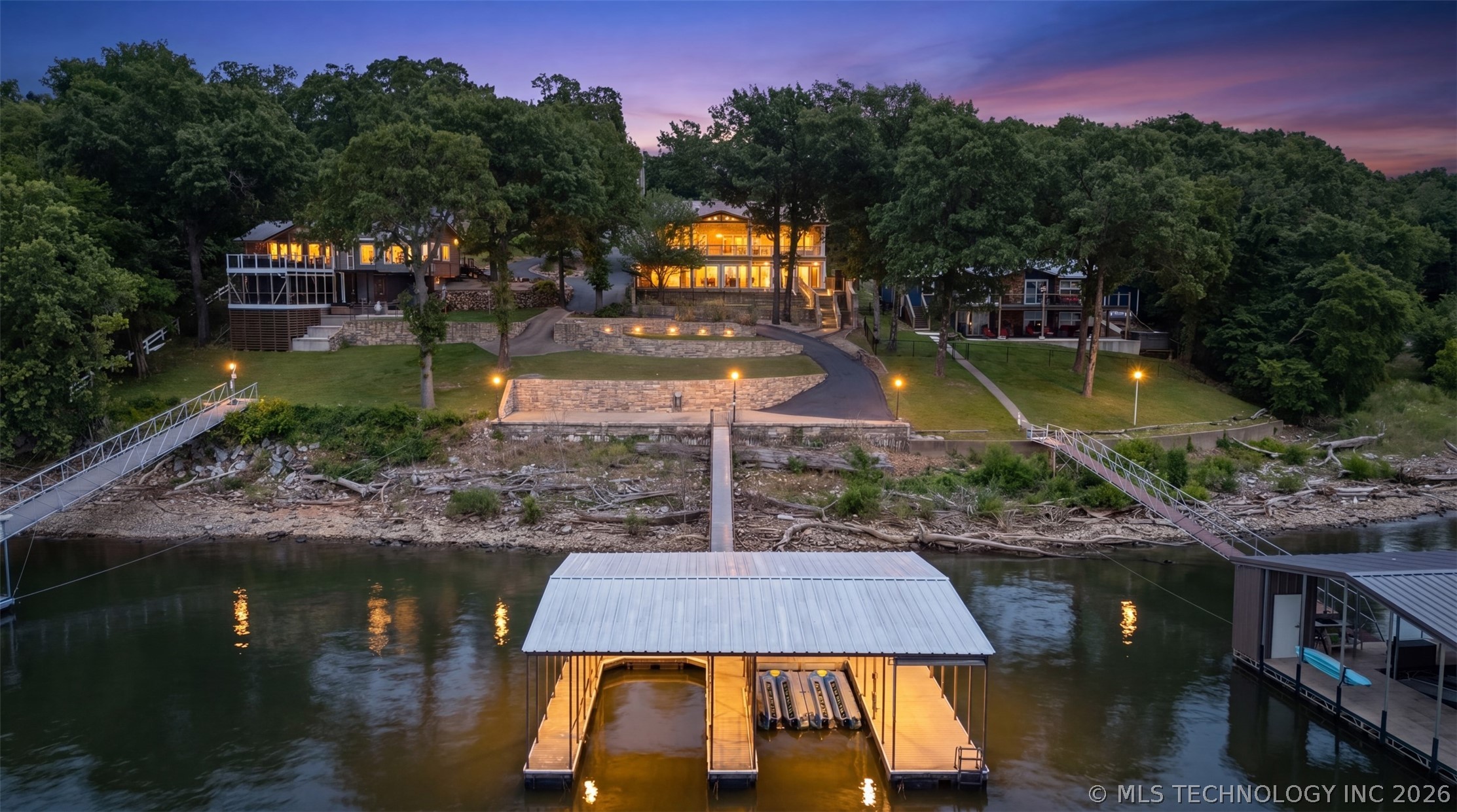 Twilight aerial of 62194 E 246th Ln, a custom waterfront home in Grove, Oklahoma, illuminated against a pink and violet sunset sky with the private dock lit on Grand Lake below